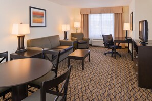 A hotel room with a sofa, two armchairs, a dining table with chairs, a TV, and a window with curtains.