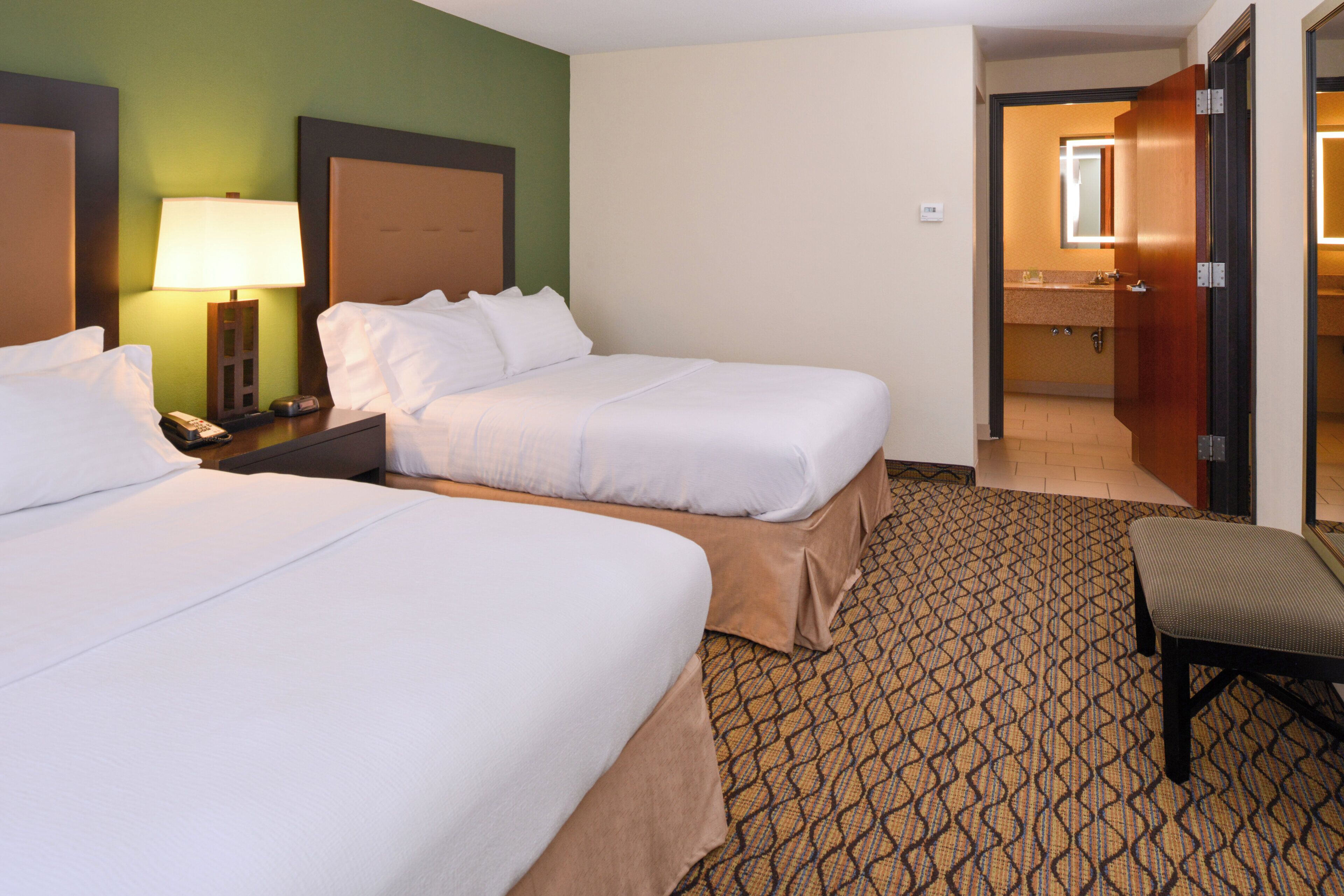 A hotel room with two beds, a nightstand, a lamp, a bathroom visible through an open door, and a patterned carpet.