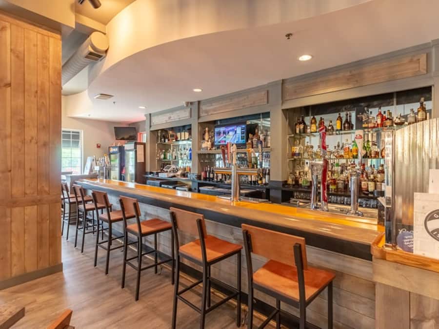 Uma área de bar com balcão extenso, vários banquinhos e variedade de garrafas e copos.