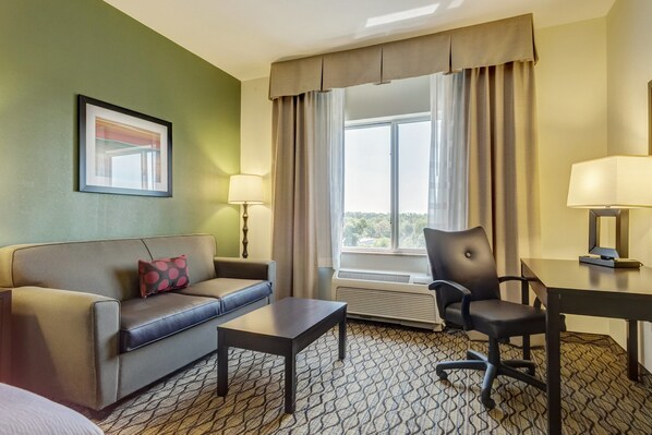 A hotel room with a sofa, a small table, a chair, a desk, two lamps, a window with curtains, and a framed picture on the wall.