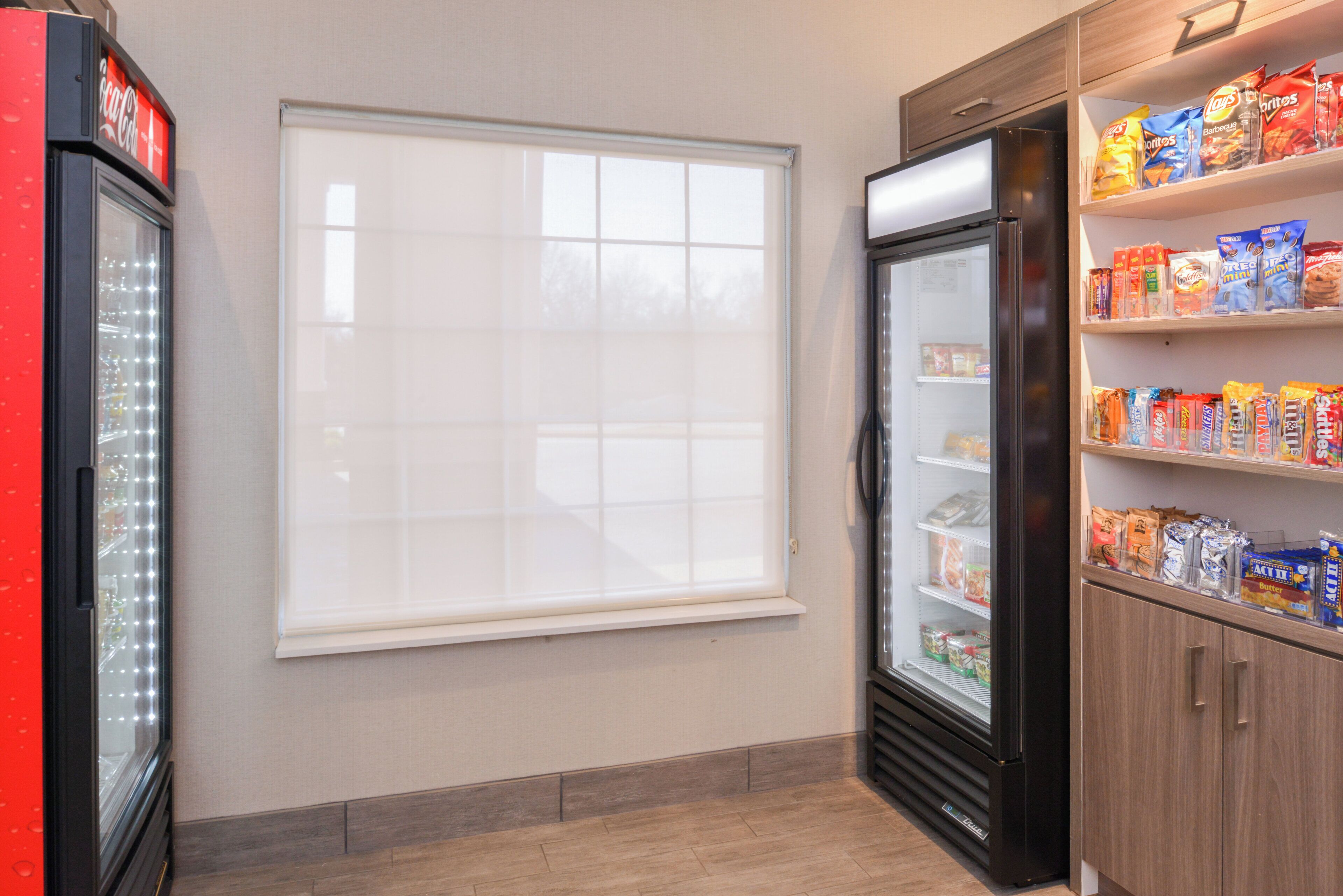 A room with a refrigerator, a window, and shelves stocked with snacks.