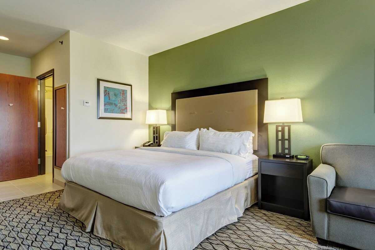 A hotel room with a bed, two bedside tables with lamps, a chair, a door, and a framed picture on the wall.