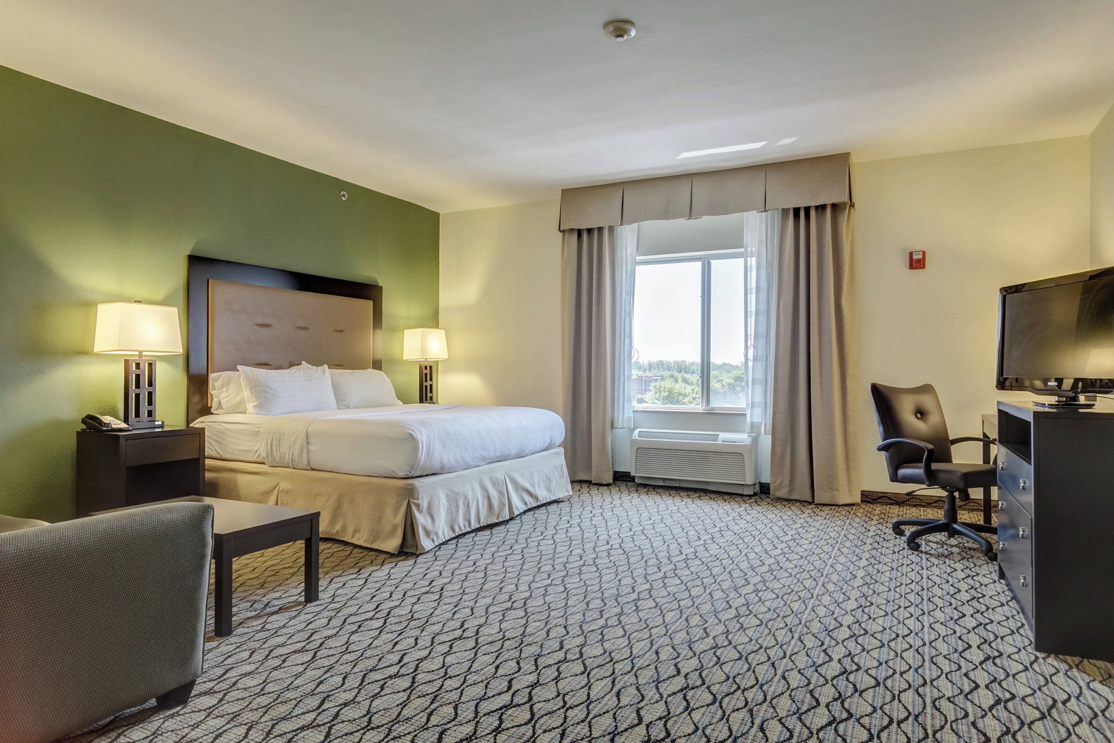 A hotel room with a bed, two bedside lamps, a desk with a chair, a TV, and a window with curtains.