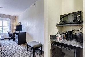A hotel room with a desk, chair, and a small seating area. It includes a microwave, coffee maker, and a refrigerator.