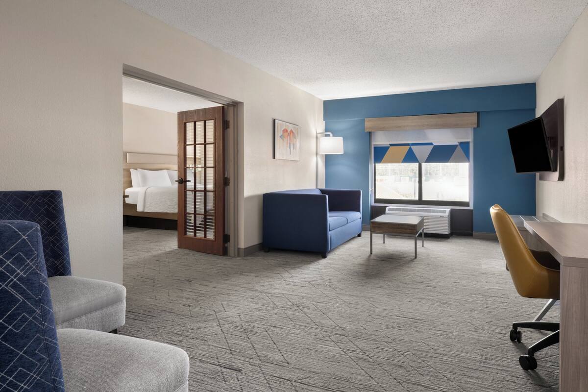 Ein Hotelzimmer mit einem Bett, einem blauen Sofa, einem Schreibtisch mit Stuhl, einem Fernseher und einem Fenster mit Jalousien.