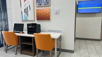 A hotel reception area with two desks, chairs, artwork on the wall, a thermostat, and a TV displaying a welcome message.