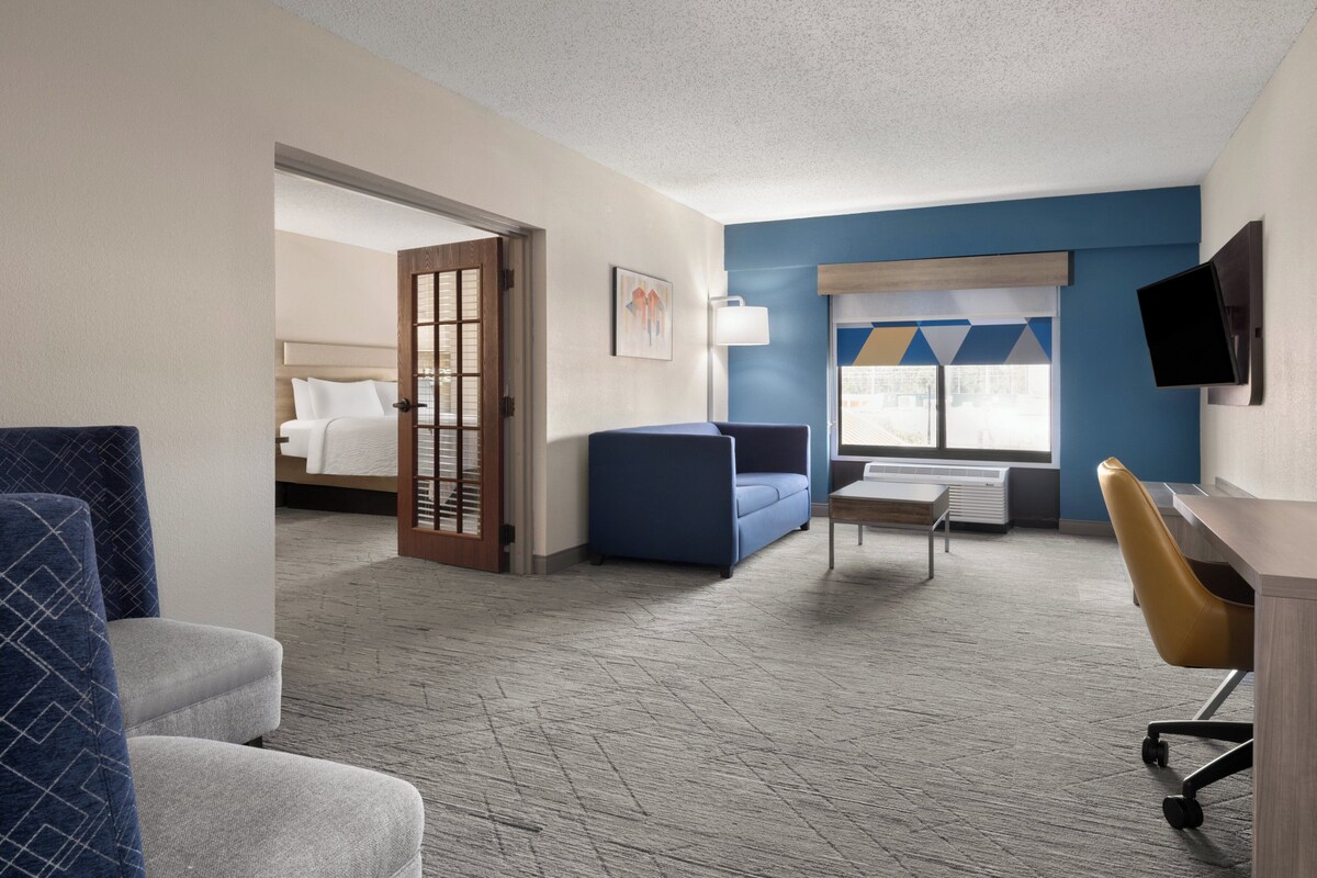 Een hotelkamer met een bed, een blauwe bank, een bureau met stoel, een televisie en een raam met zonwering.
