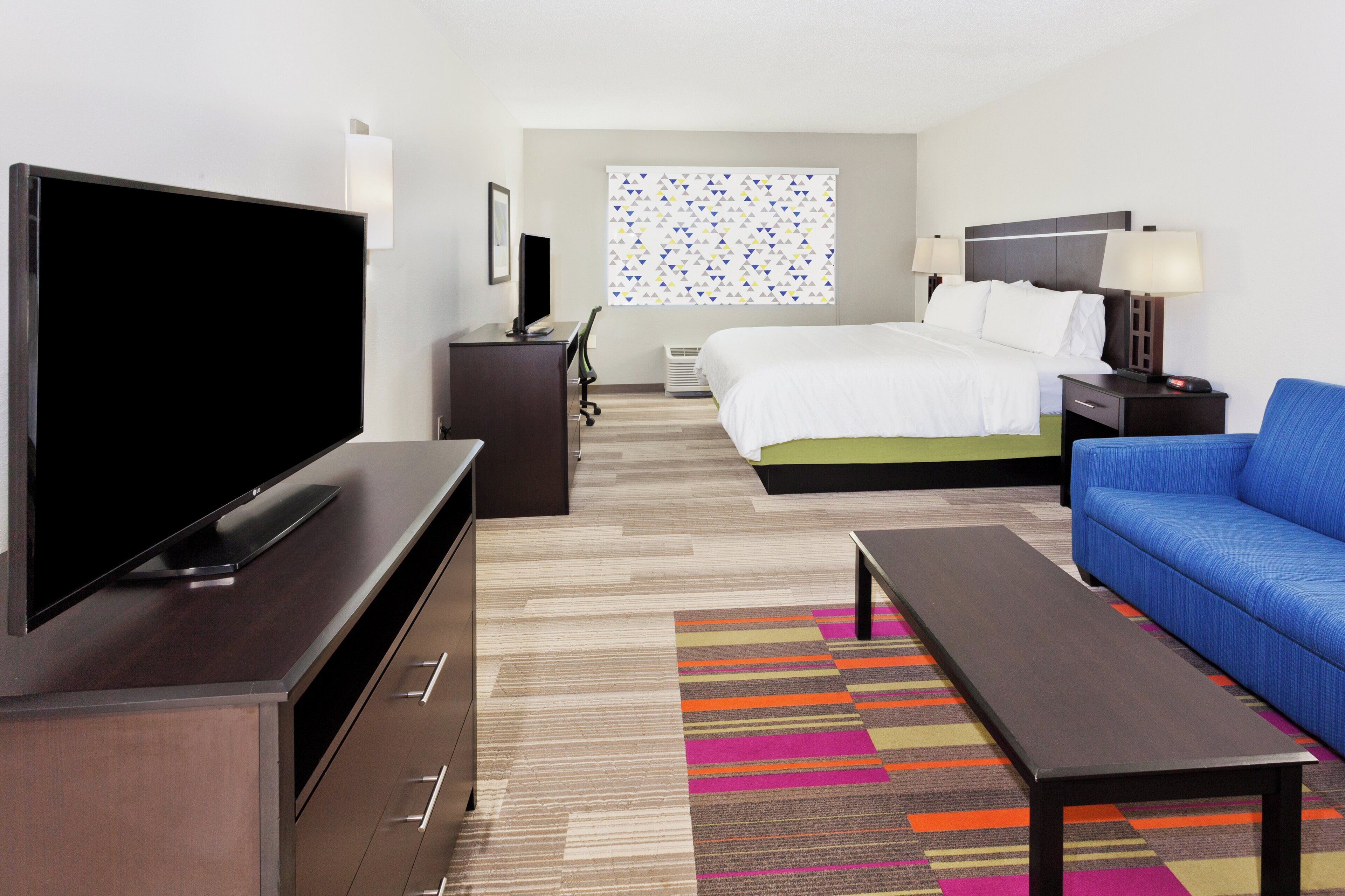 A hotel room with a television, a bed, a desk, a chair, a sofa, and a colorful rug.
