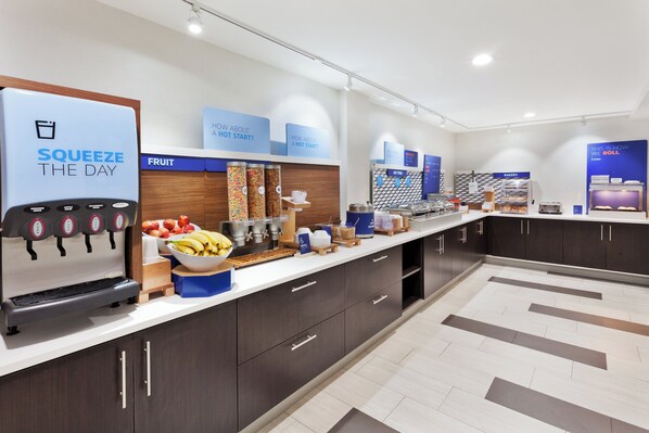 A modern breakfast bar with a juice dispenser, fruit, and hot food options.