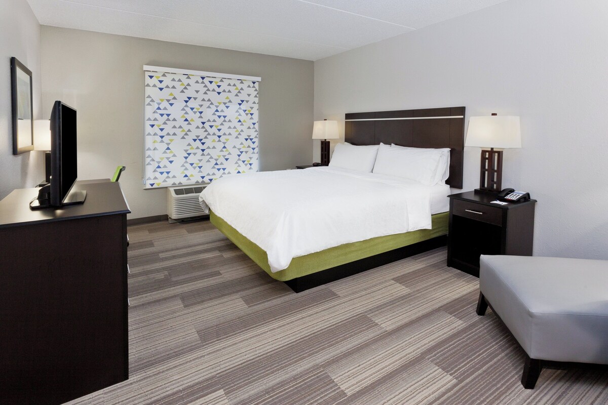 Une chambre d’hôtel comprenant un lit, des tables de chevet, un bureau, une chaise, une fenêtre avec des stores et un appareil de climatisation.