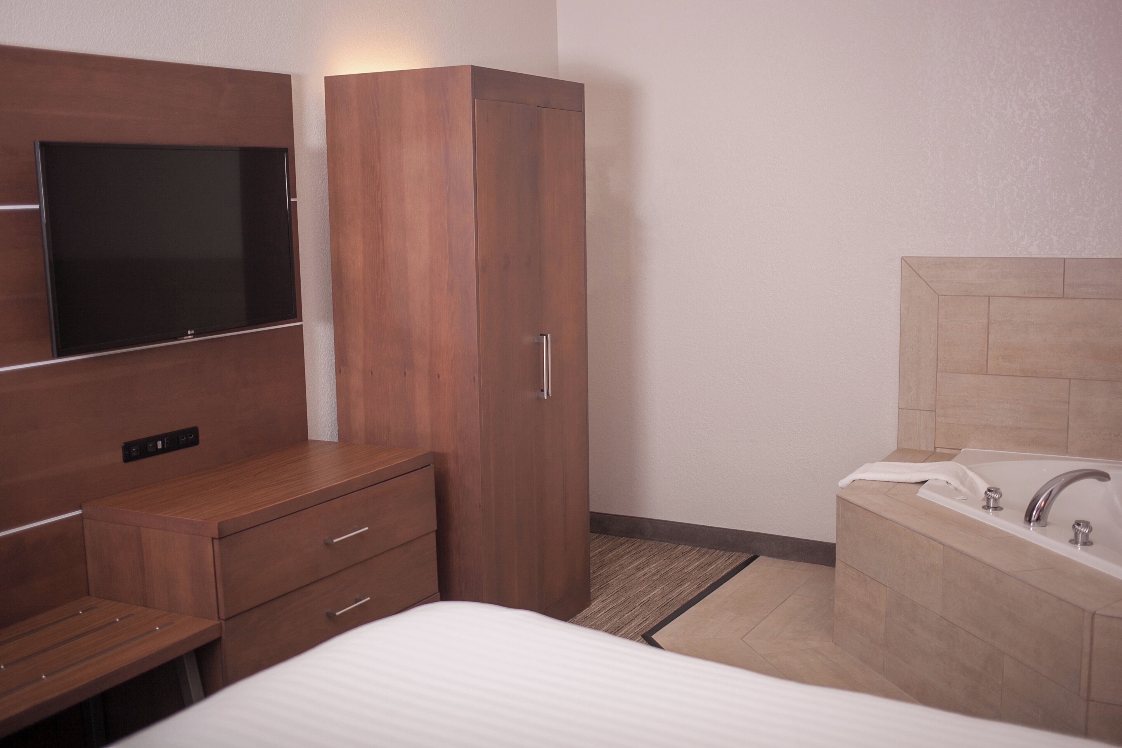 A hotel room with a bed, a wooden wardrobe, a TV mounted on the wall, and a bathroom with a bathtub and sink.