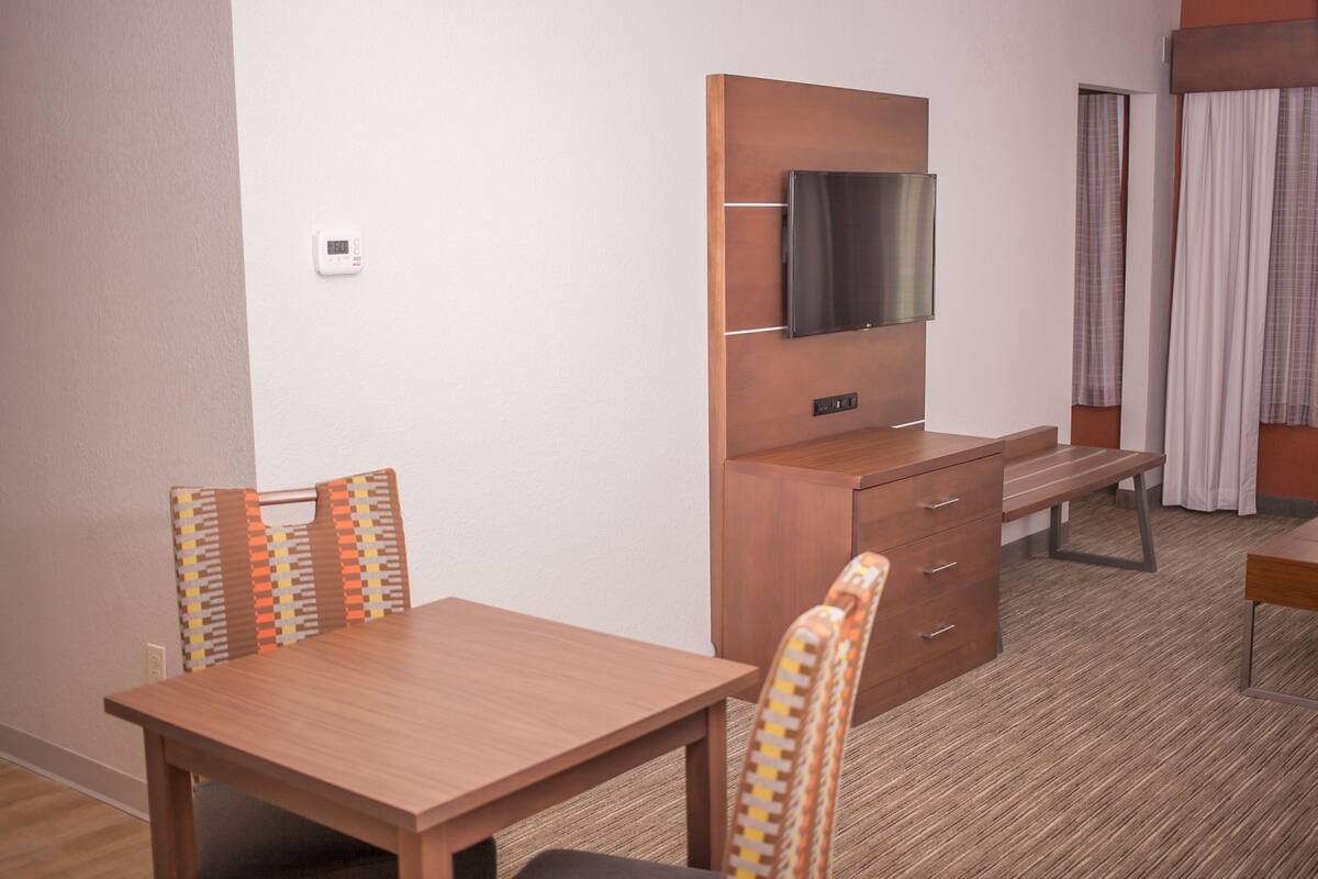 Ett hotellrum med ett matbord i trä, två stolar, en tv som är monterad på ett träskåp och ett litet skrivbord med lådor.
