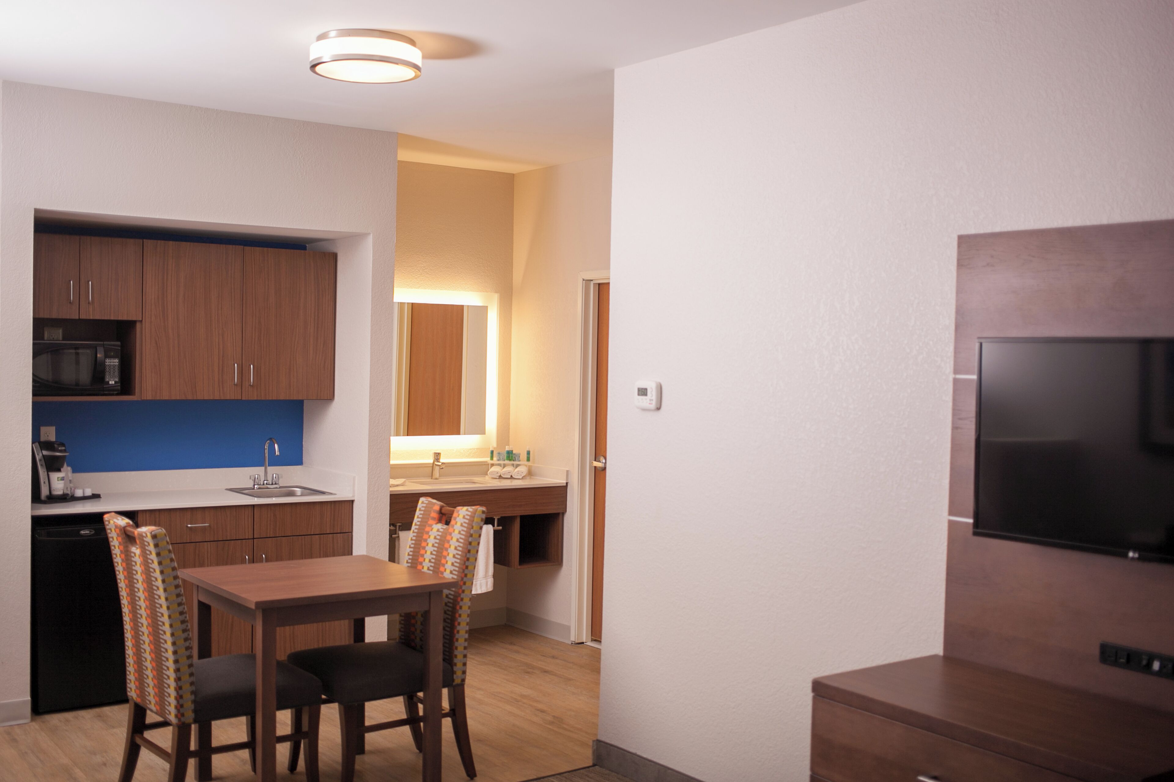A compact kitchen with wooden cabinets, a microwave, and a sink. There is a dining area with a table and chairs. A flat-screen TV is mounted on the wall.