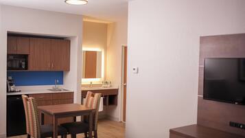 A compact kitchen with wooden cabinets, a microwave, and a sink. There is a dining area with a table and chairs. A flat-screen TV is mounted on the wall.