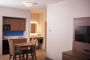 Cocina funcional con muebles de madera, microondas y lavamanos. Hay una zona de comedor con mesa y sillas. Un televisor de pantalla plana está montado en la pared.