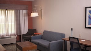 A modern hotel room with a sofa, a small table, a desk, a chair, a floor lamp, and a large window with curtains. - Holiday Inn Express Hotel & Suites by IHG (Logan)