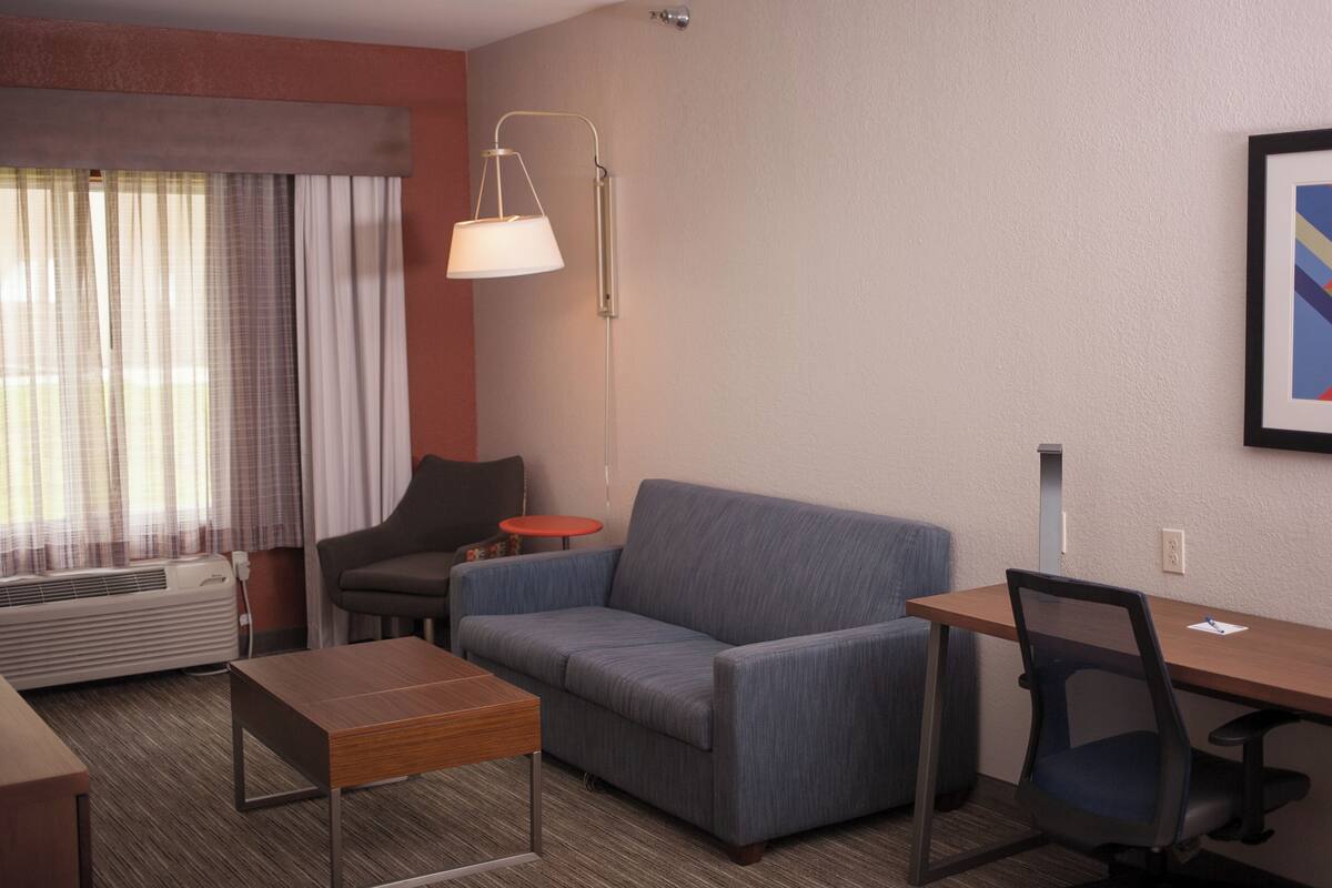 Ett modernt hotellrum med en soffa, ett litet bord, ett skrivbord, en stol, en golvlampa och ett stort fönster med gardiner.