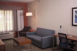 Habitación de hotel con sofá, una mesita, escritorio, silla, lámpara y ventana con cortinas.