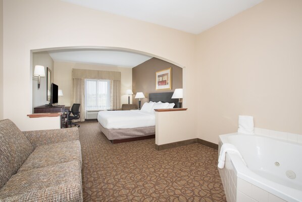 A hotel room with a bed, a sofa, a desk, a chair, a desk lamp, a wall-mounted TV, a framed picture, and a bathtub. - Holiday Inn Express Hotel & Suites Concordia by IHG (Concordia)
