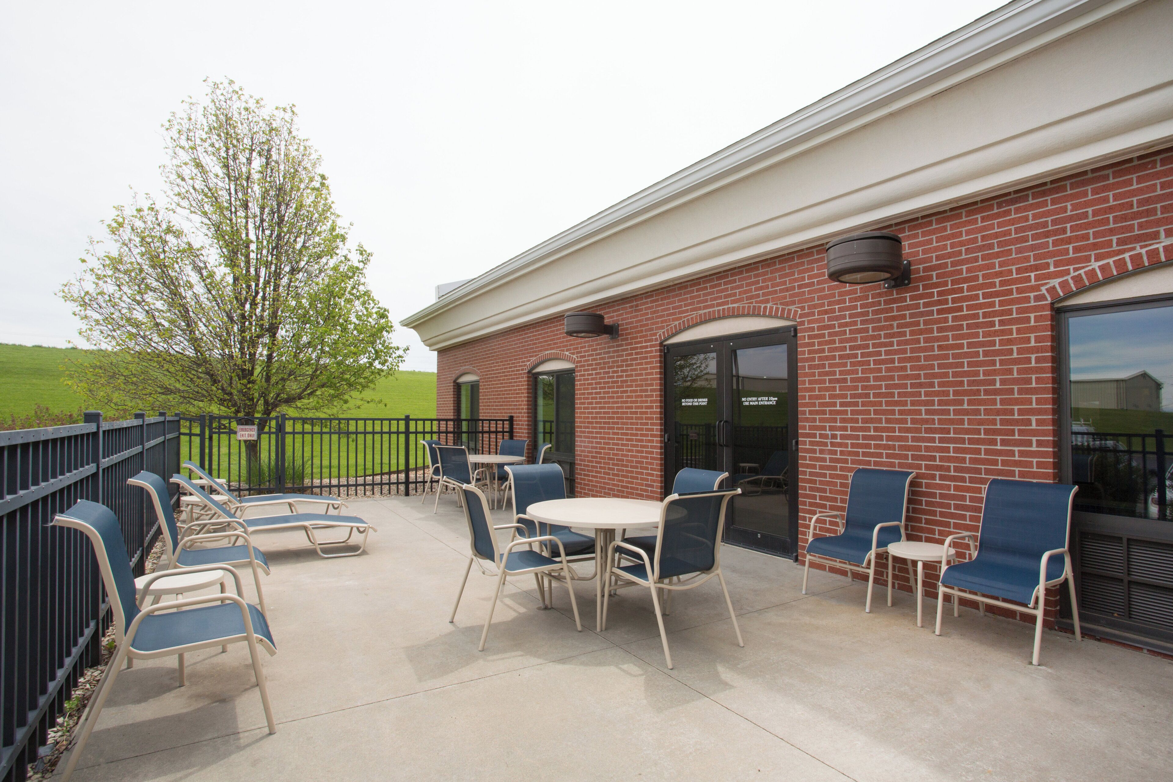 An outdoor patio with tables and chairs, a brick building, and a green field in the background.