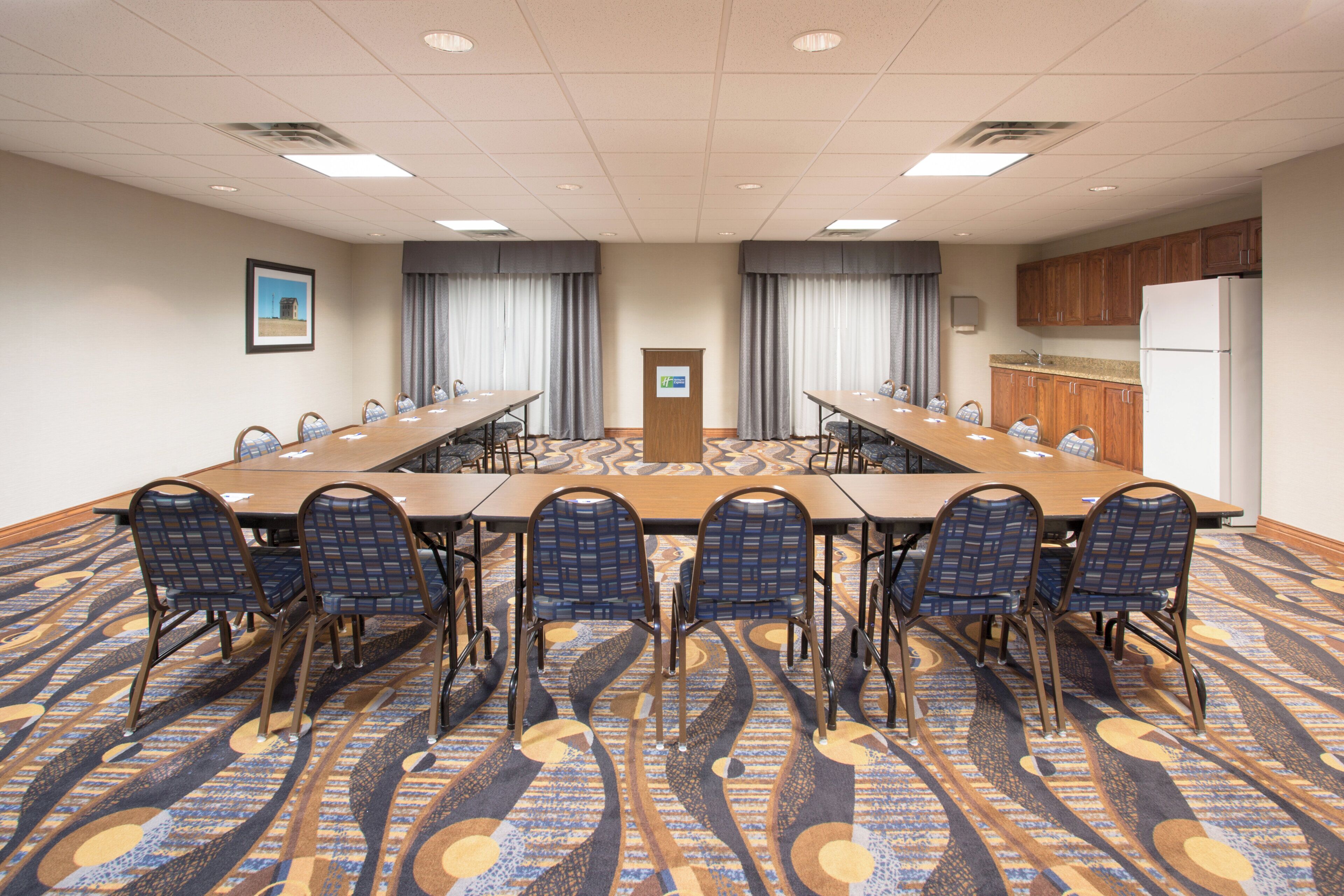 A conference room with a long table, chairs, a podium, curtains, and a kitchen area.
