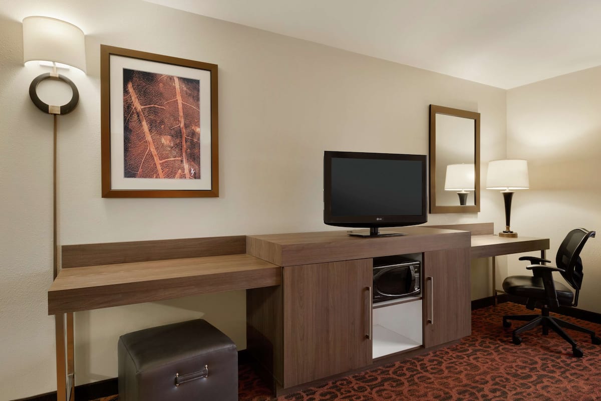 Habitación de hotel con escritorio de madera, televisor, silla, refrigerador pequeño, maleta, un cuadro enmarcado y un espejo.