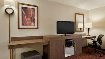 A hotel room with a wooden desk, a television, a chair, a small refrigerator, a suitcase, a framed picture, and a mirror.