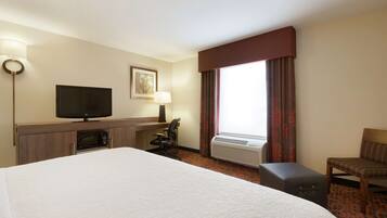 Quarto de hotel com uma cama grande, uma escrivaninha com televisão de tela plana, uma cadeira, aparelho de ar condicionado e uma janela com cortinas.