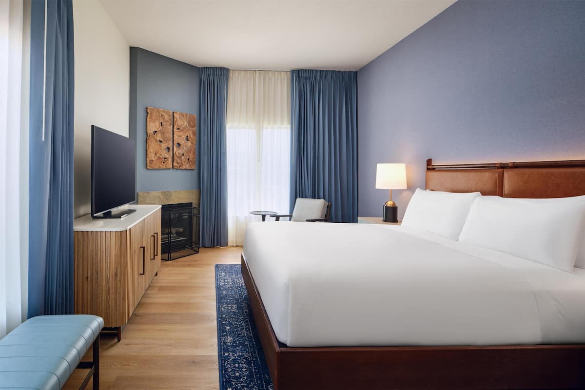 A hotel room with a bed, a desk with a TV, a chair, a lamp, and artwork on the wall.