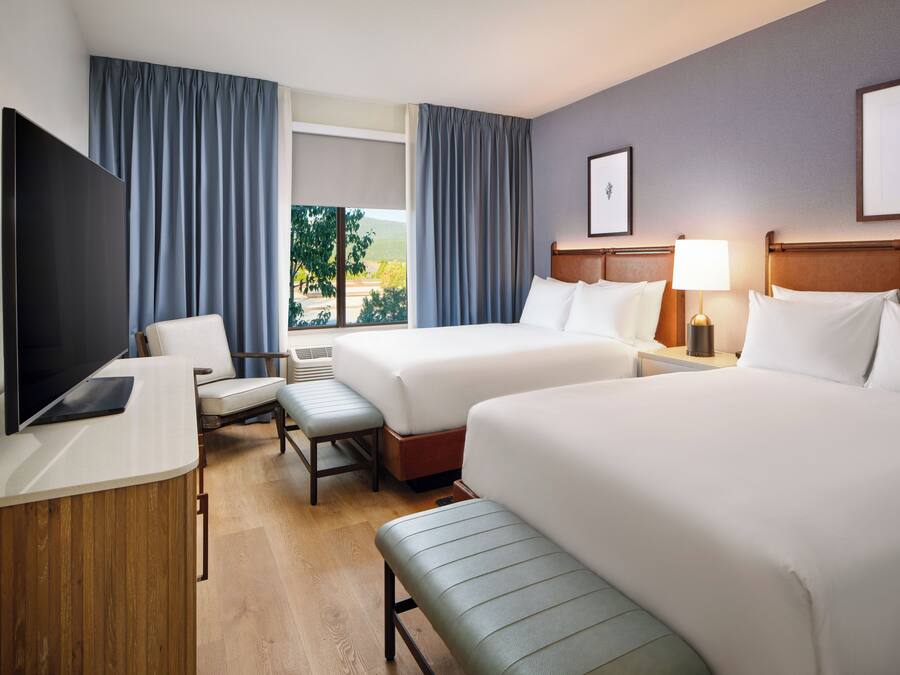 Hotelkamer met twee bedden, een bureau, een flatscreen televisie, een stoel, een bankje en een raam met gordijnen.