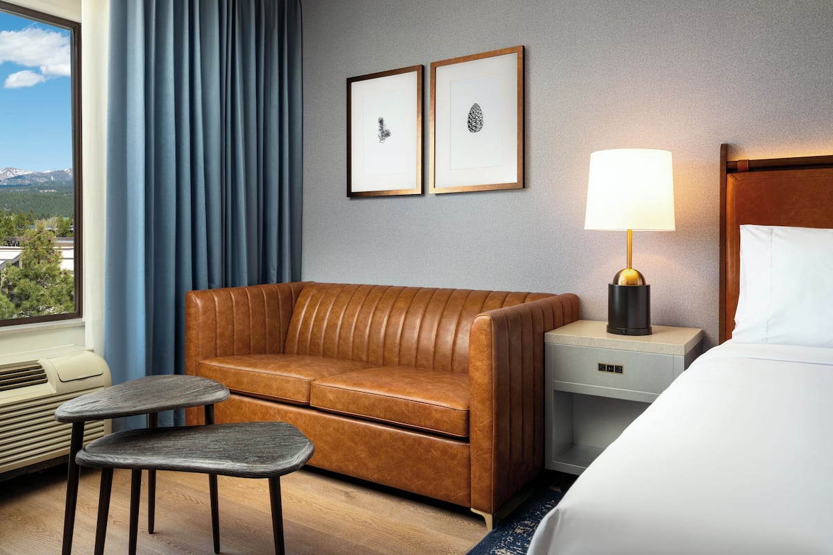 A hotel room with a brown leather sofa, a small table, a bedside lamp, and a bed. There are two framed pictures on the wall and a window with curtains.