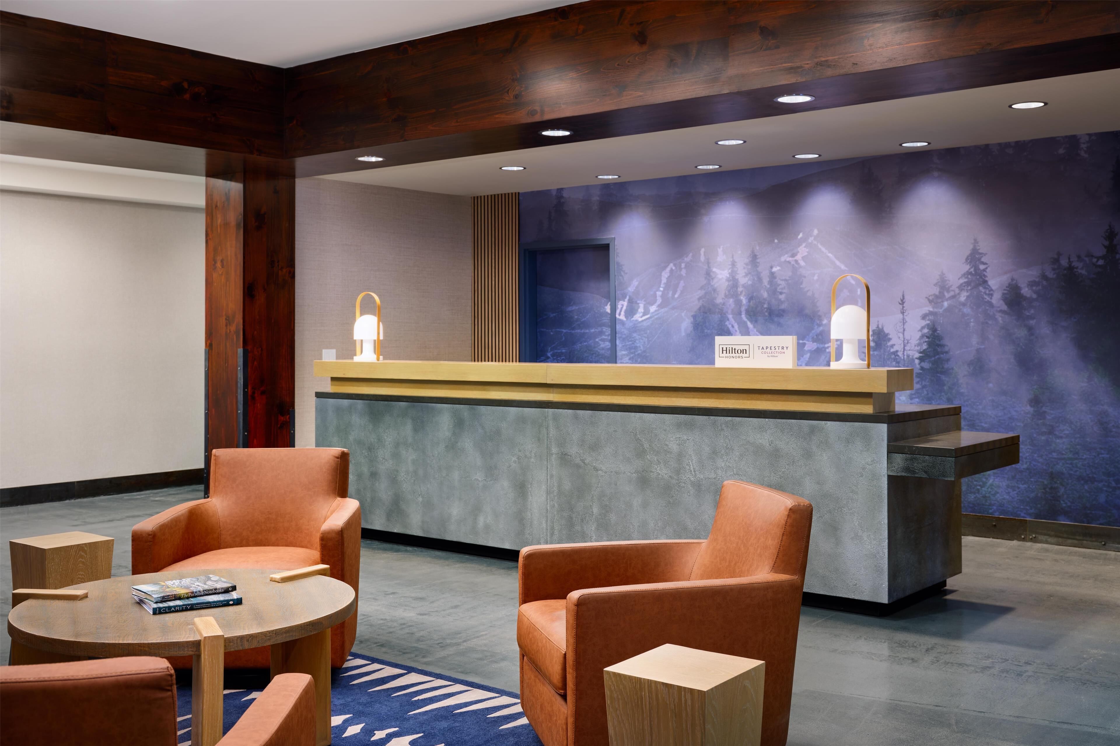 A reception area with a wooden reception desk, two orange chairs, a round table with magazines, and a blue carpet.