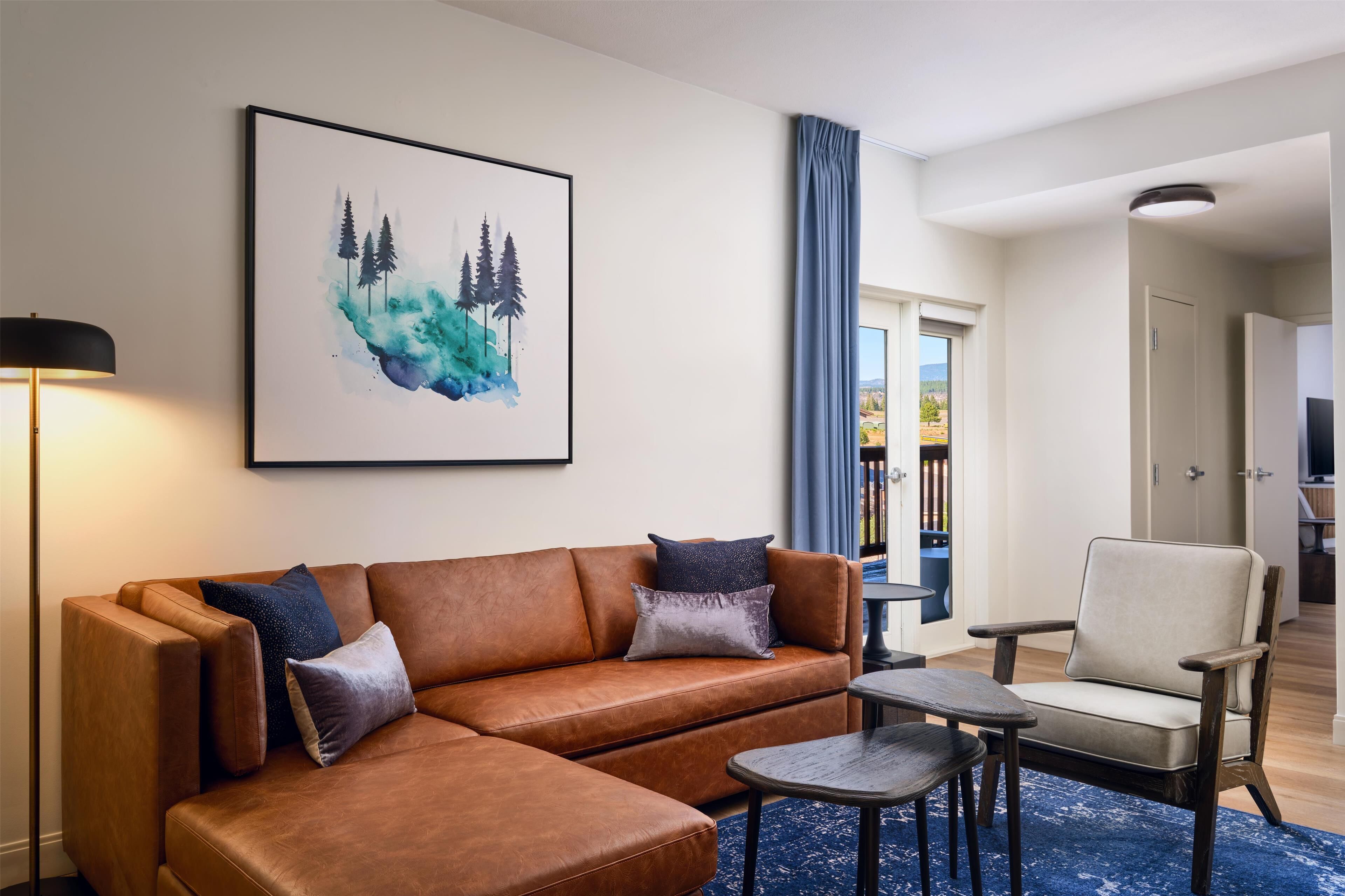 A living room with a brown leather sofa, a chair, a small table, and a painting of trees on the wall.