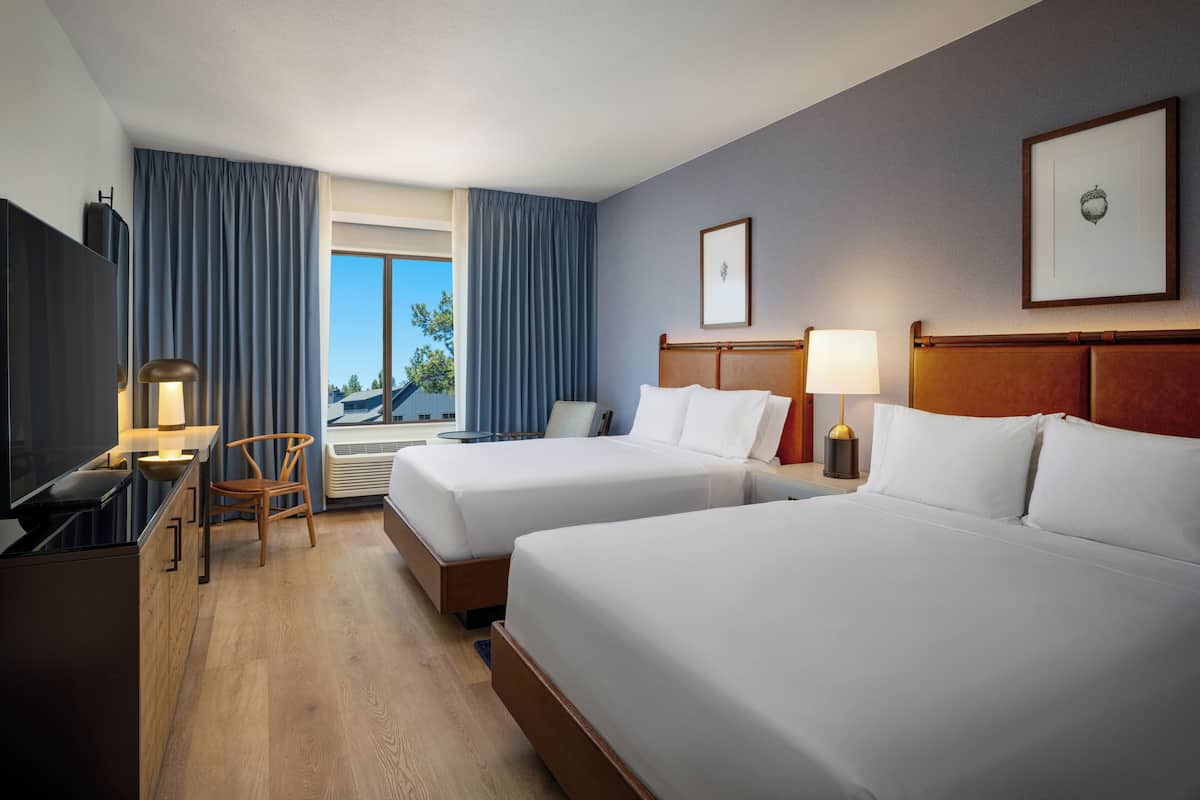 A hotel room with two beds, a desk, a chair, a lamp, and a window with curtains.