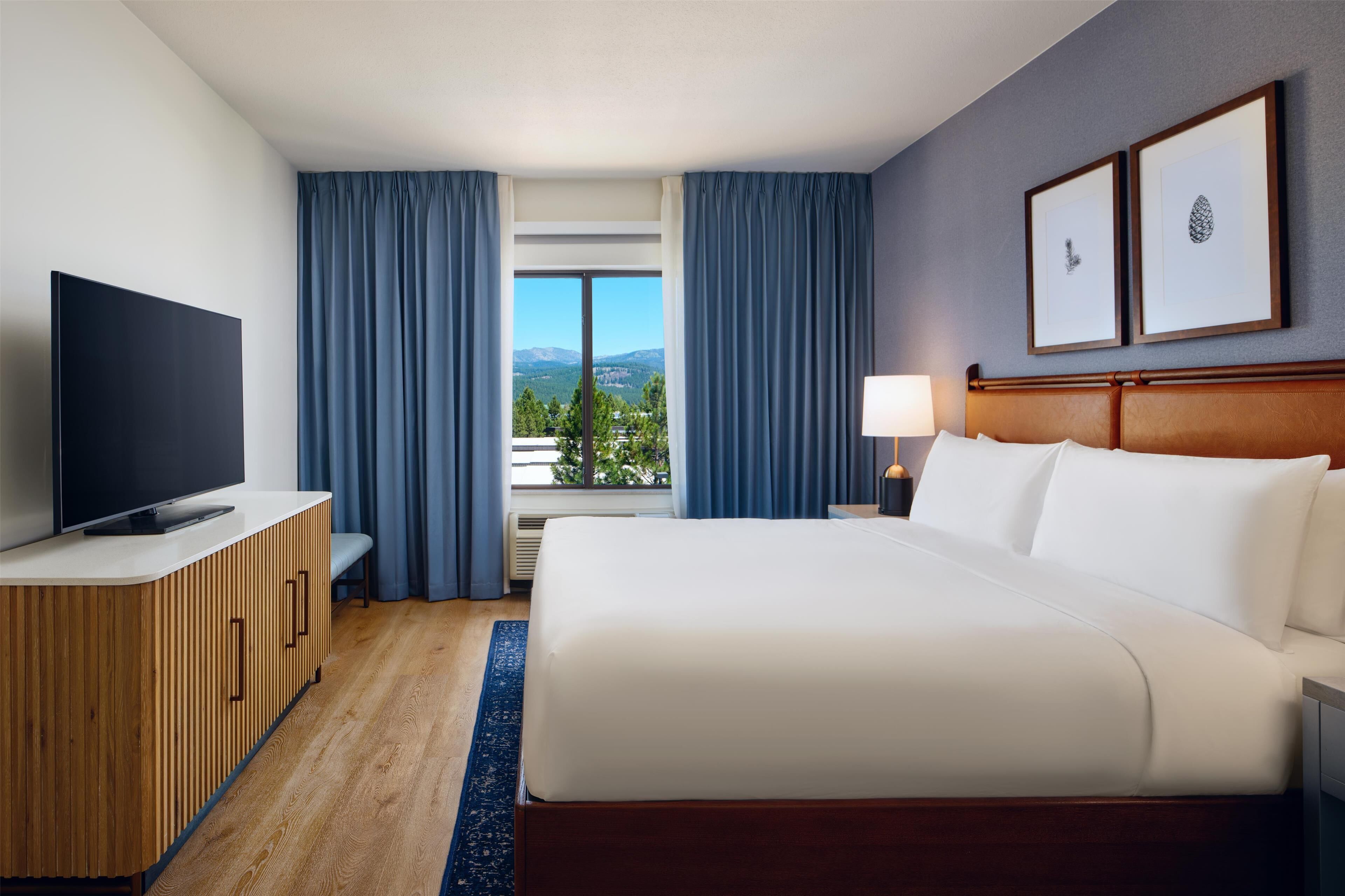 A hotel room with a bed, a TV, a desk, a lamp, and a window with curtains.