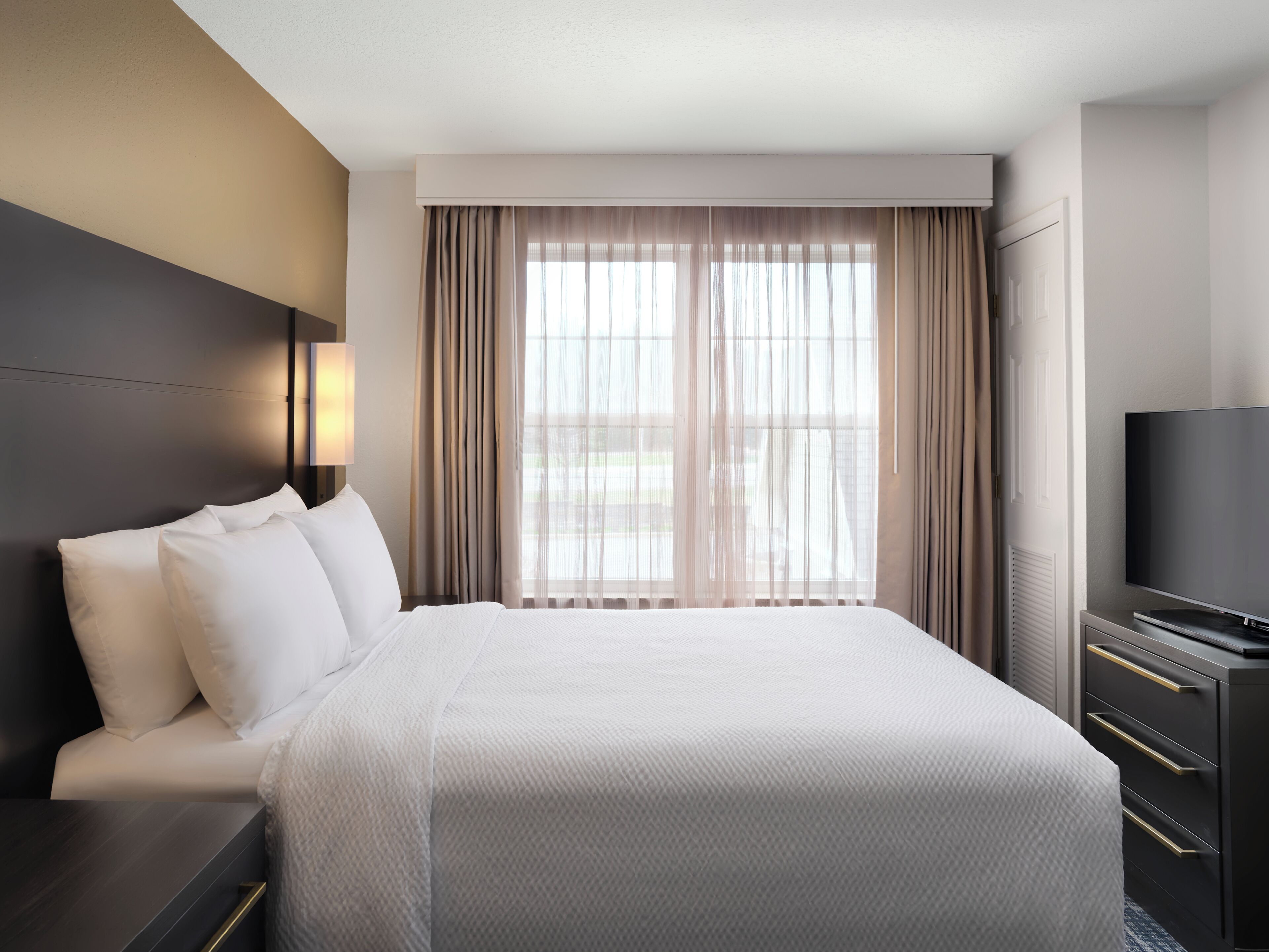 A hotel room with a large bed, a TV, and a window with sheer curtains.