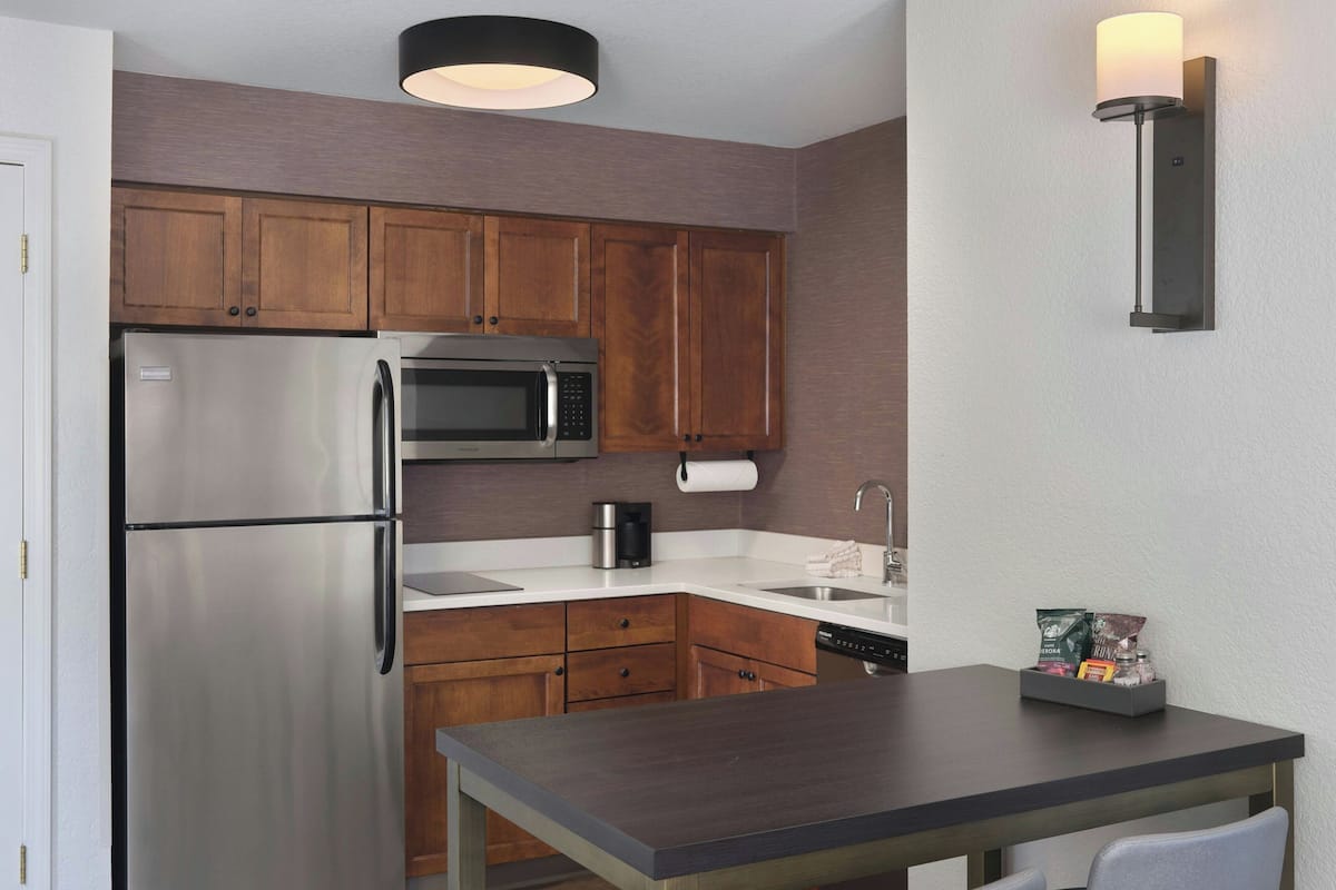Una cocina compacta con armarios de madera, un refrigerador de acero inoxidable, un microondas, una cafetera y una pequeña mesa con una silla.