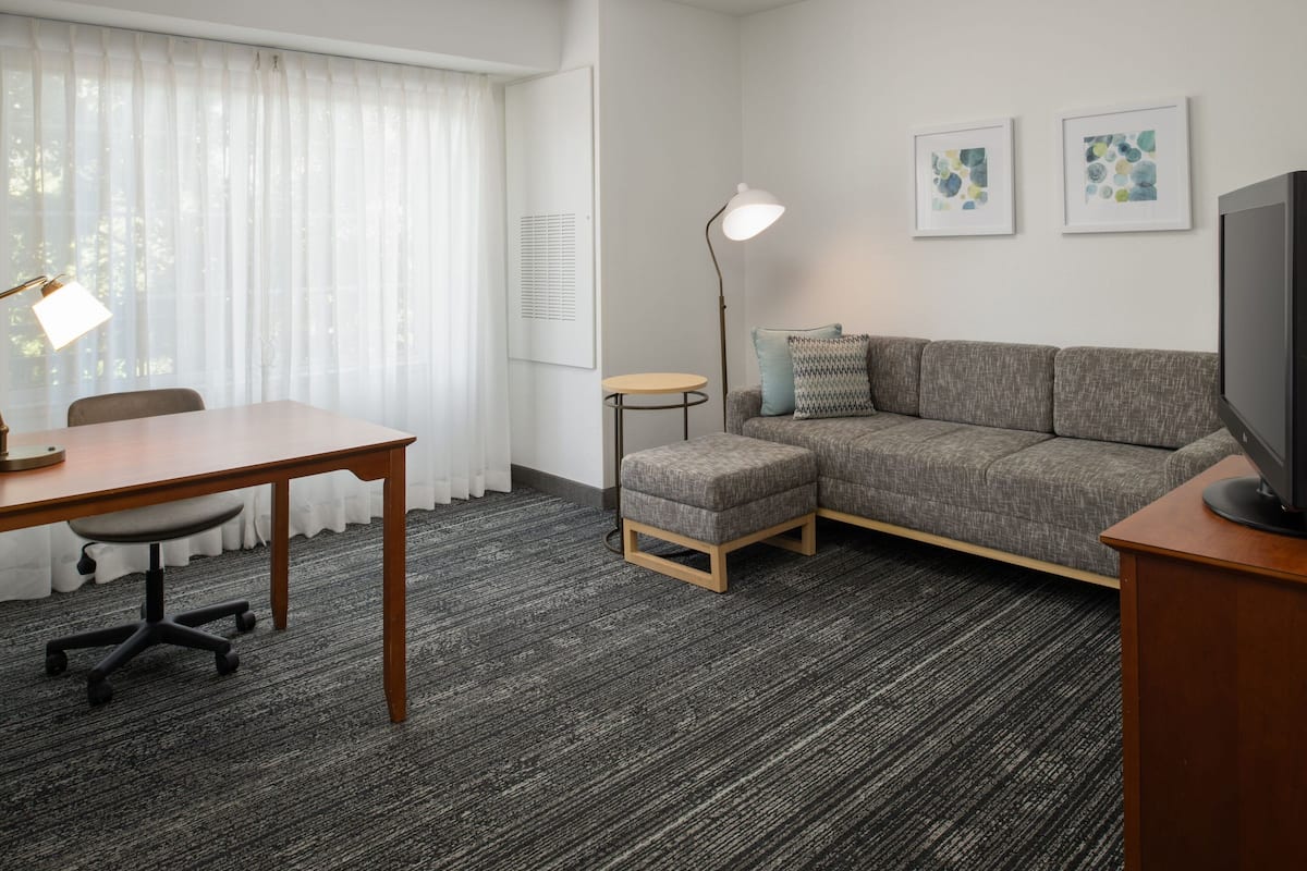 Una habitación de hotel moderna con alfombra gris, un sofá gris, una pequeña mesa redonda, un escritorio con una silla, una lámpara de pie y un televisor sobre un soporte de madera.