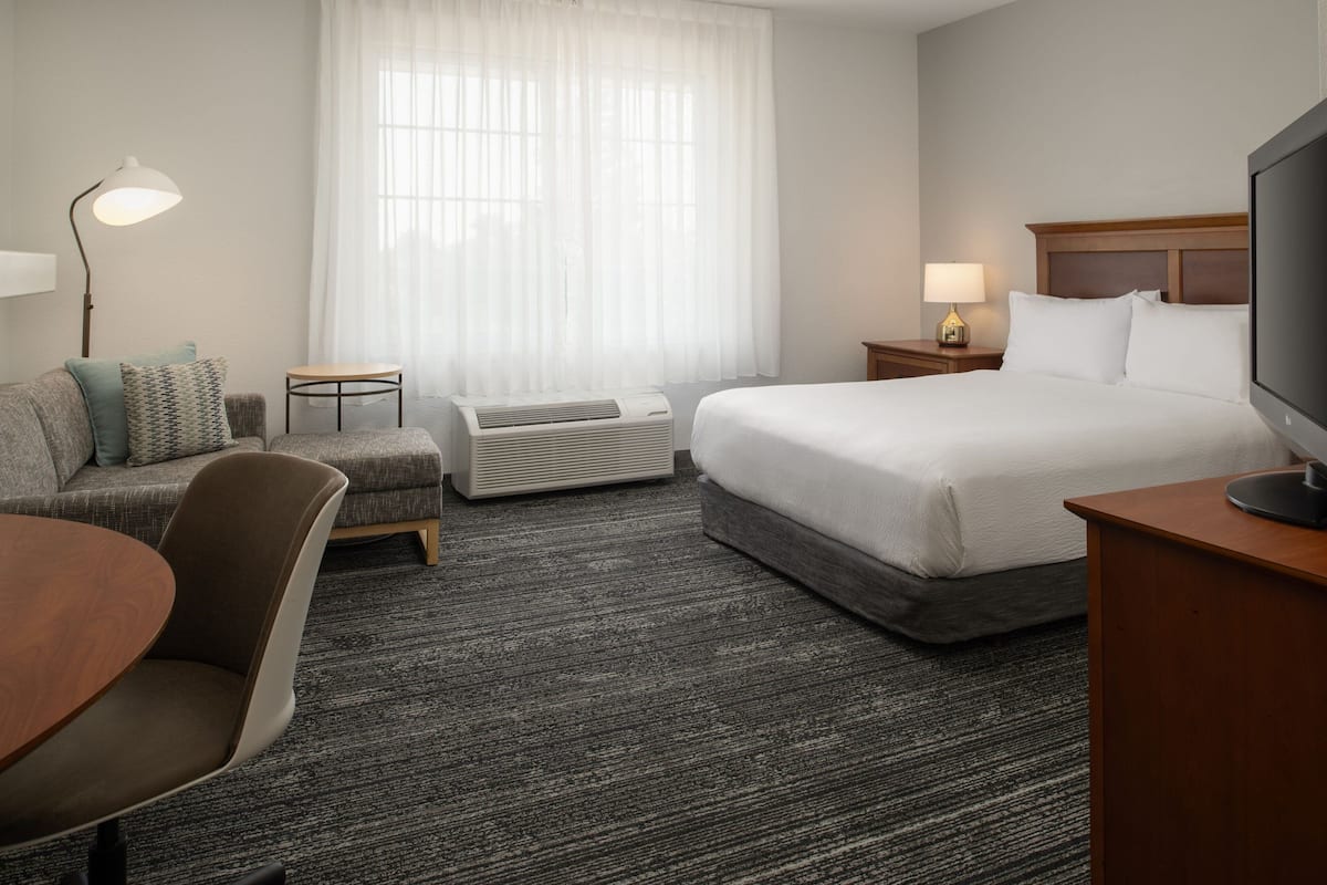 Una habitación de hotel con cama, escritorio, silla, lámpara y ventana con cortinas translúcidas.