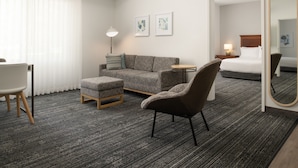 Une chambre d’hôtel avec un canapé gris, une petite table ronde, une chaise, un lit et un grand miroir.