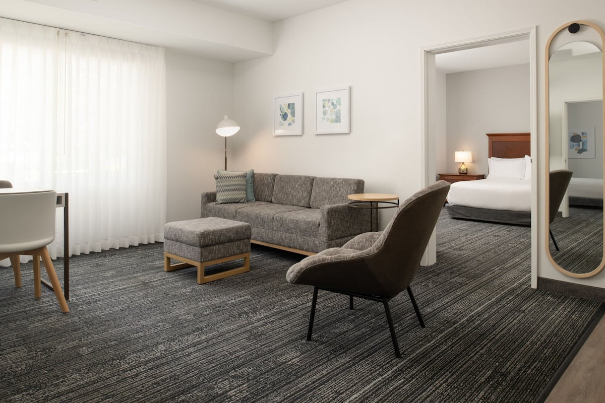 Habitación de hotel con sofá gris, una mesita redonda pequeña, una silla, una cama y un espejo grande.
