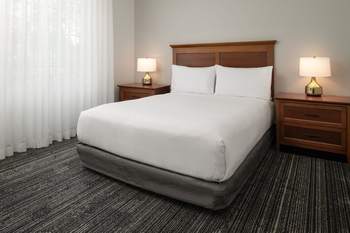 Habitación de hotel con una cama grande, dos mesitas de noche con lámparas, un cabecero de madera y suelo alfombrado.