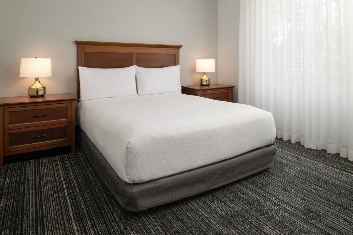 Habitación de hotel con una cama grande, dos mesitas de noche con lámparas, cabecero de madera, piso alfombrado y una ventana con cortinas traslúcidas.