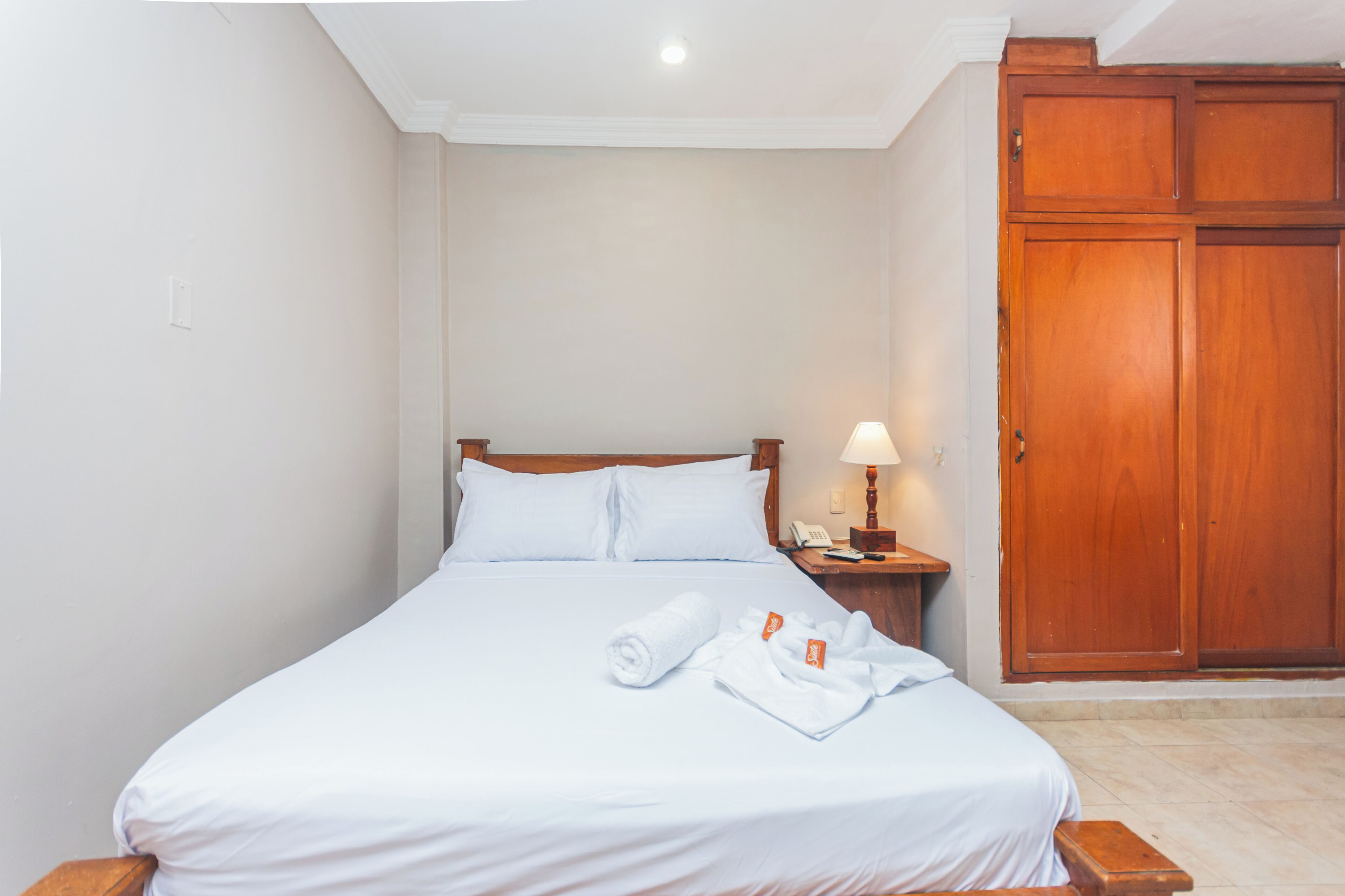 A neatly made bed with white linens, a wooden nightstand with a lamp, and a wooden wardrobe.