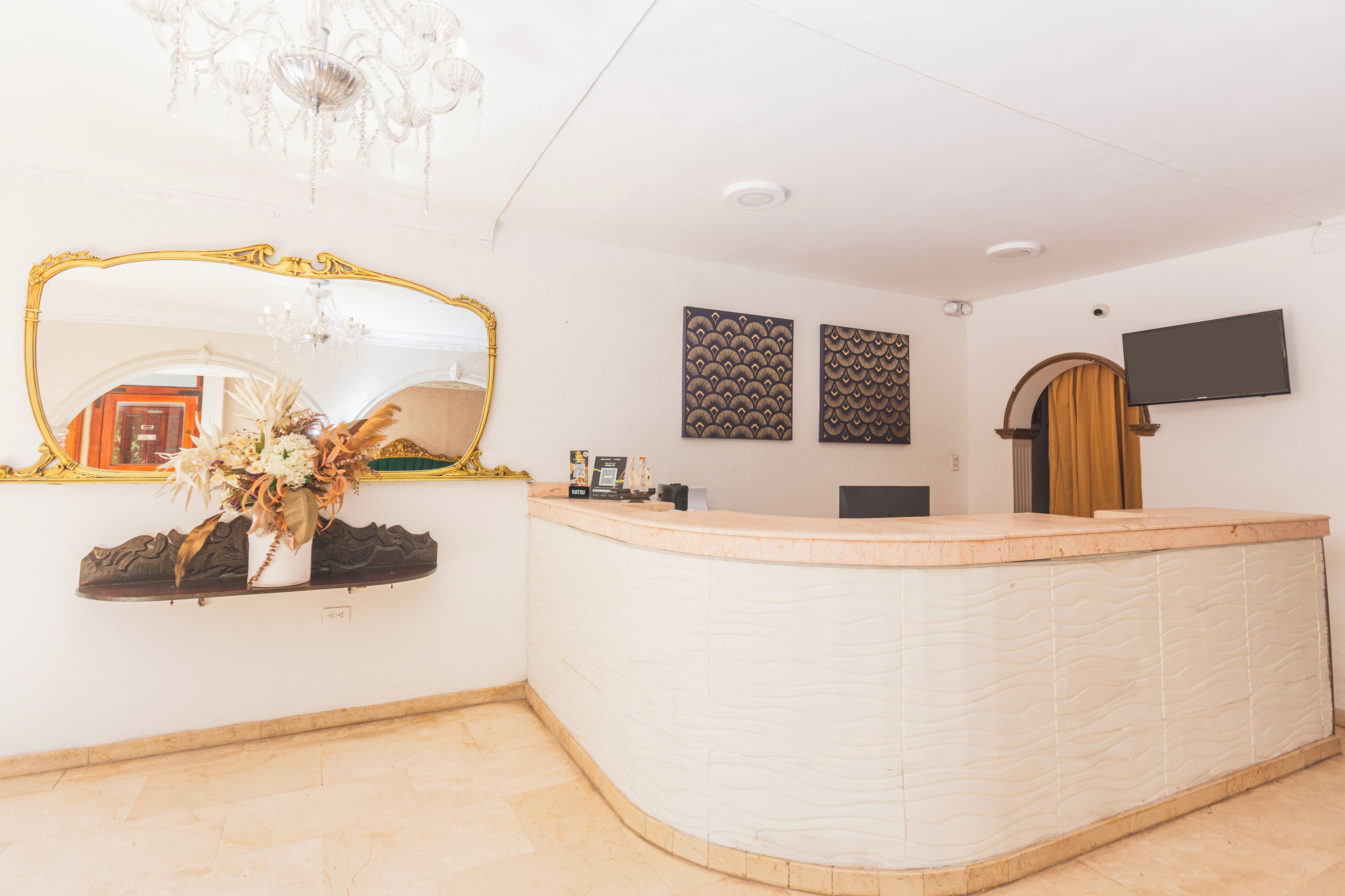A reception area with a curved reception desk, a large mirror with an ornate frame, a chandelier, and two decorative wall panels.