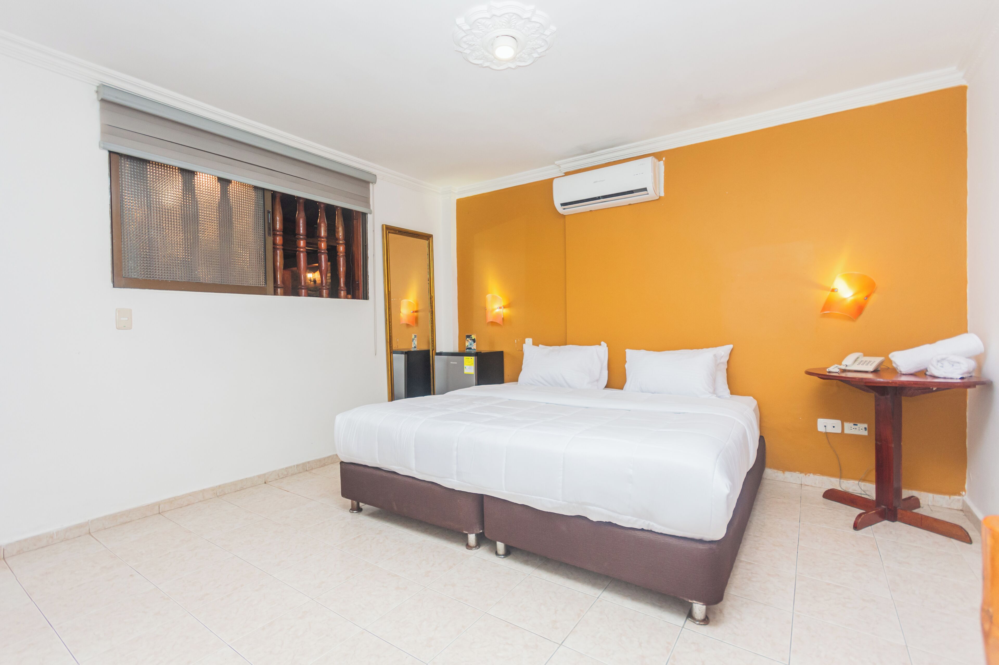 Quarto de hotel com uma cama grande, uma mesinha com telefone e toalha, uma janela com persianas e um ar condicionado na parede.