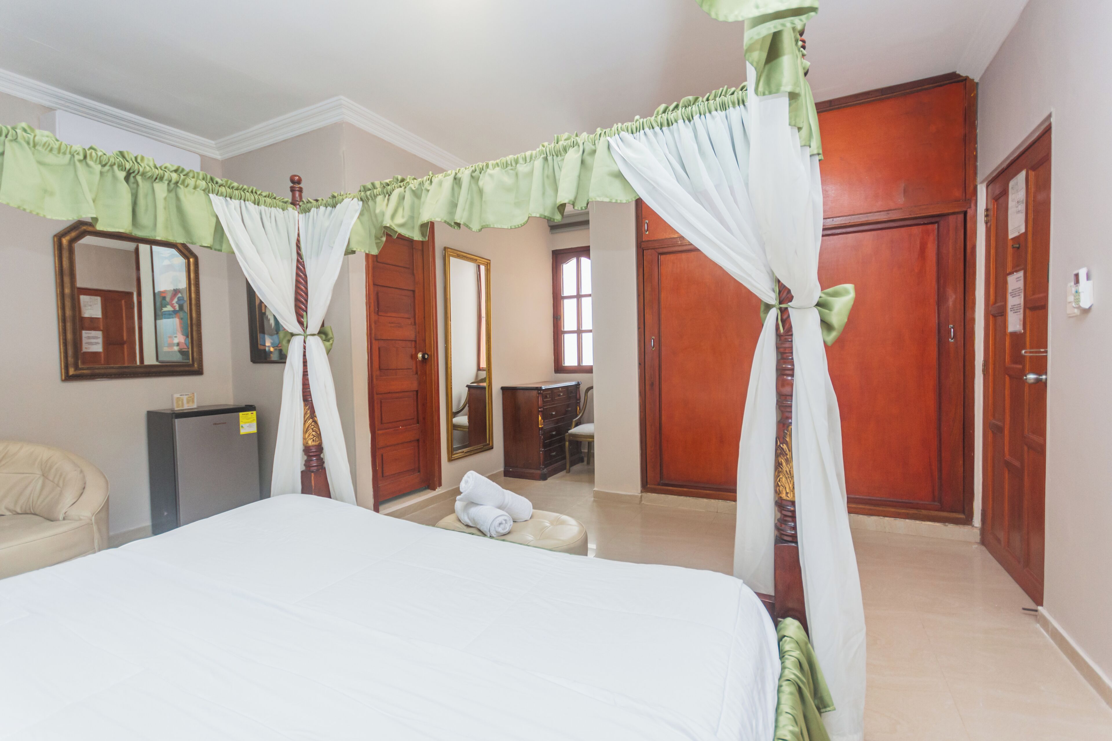 A bedroom with a four-poster bed, a small desk, a chair, a wardrobe, and a mirror.