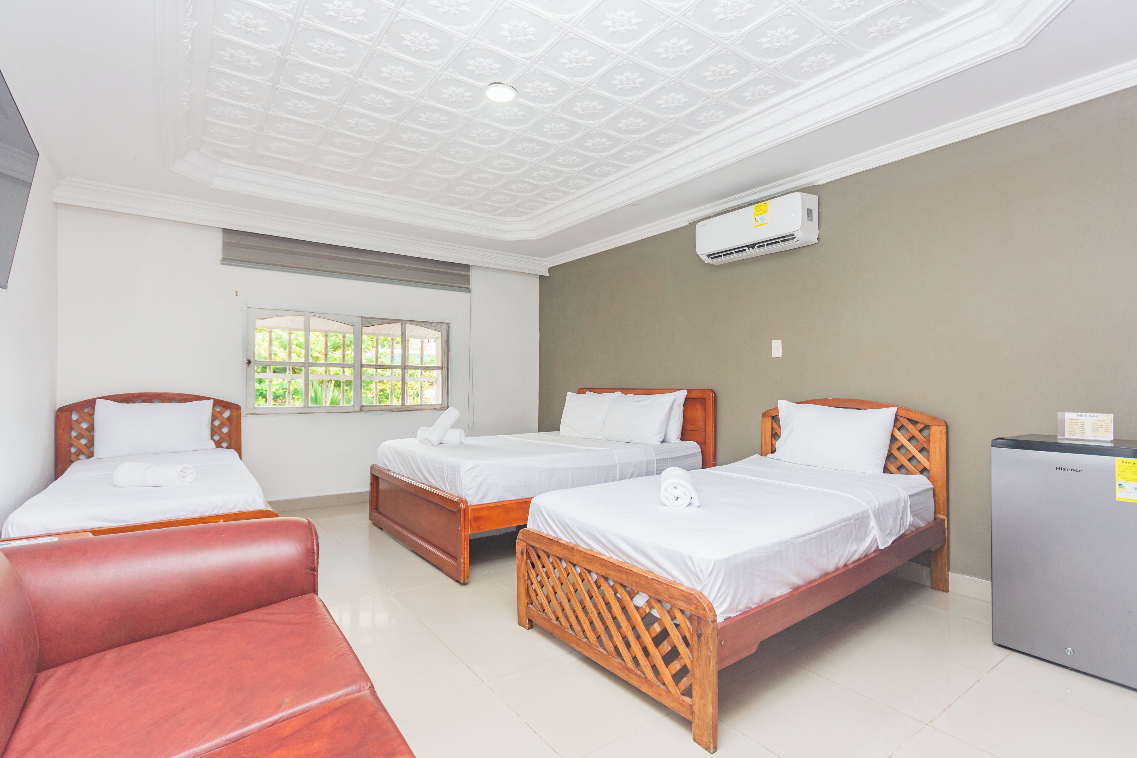 A hotel room with two wooden beds, a red sofa, a small refrigerator, and a window with a view of greenery.
