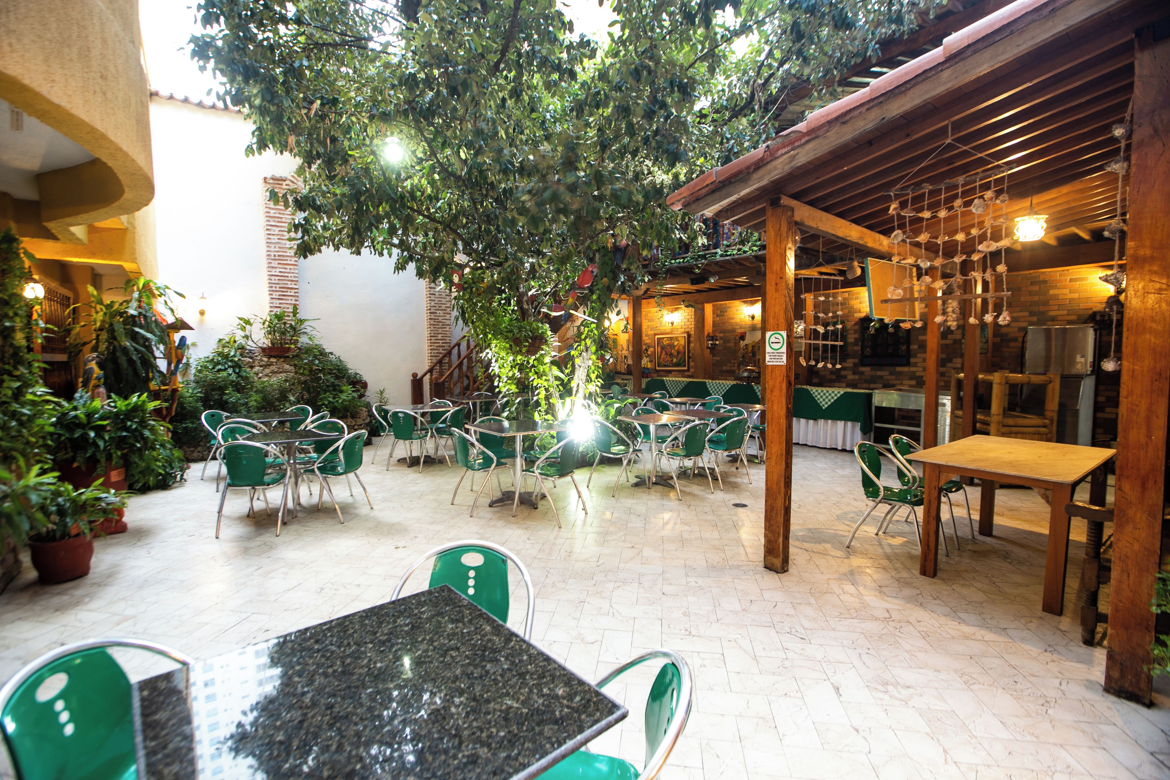 An outdoor patio area with green metal chairs and tables, surrounded by potted plants and a large tree.