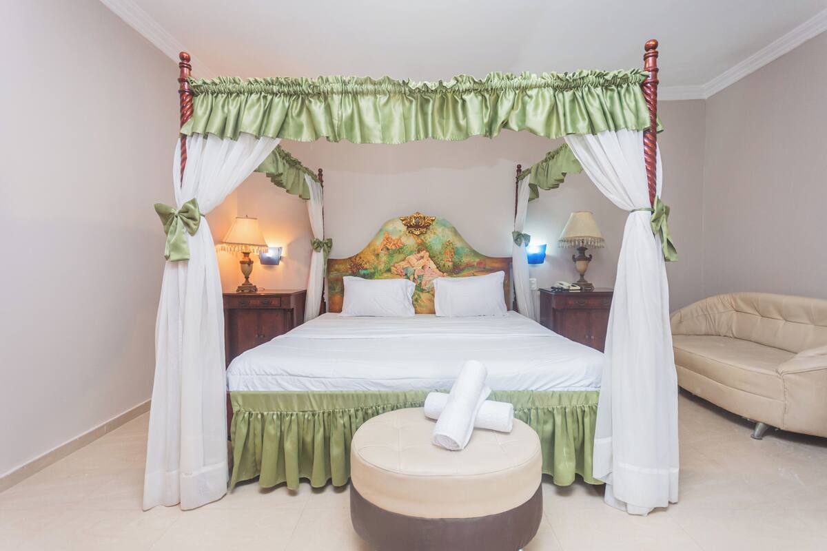 A four-poster canopy bed with a floral headboard, a round ottoman, and a beige sofa.
